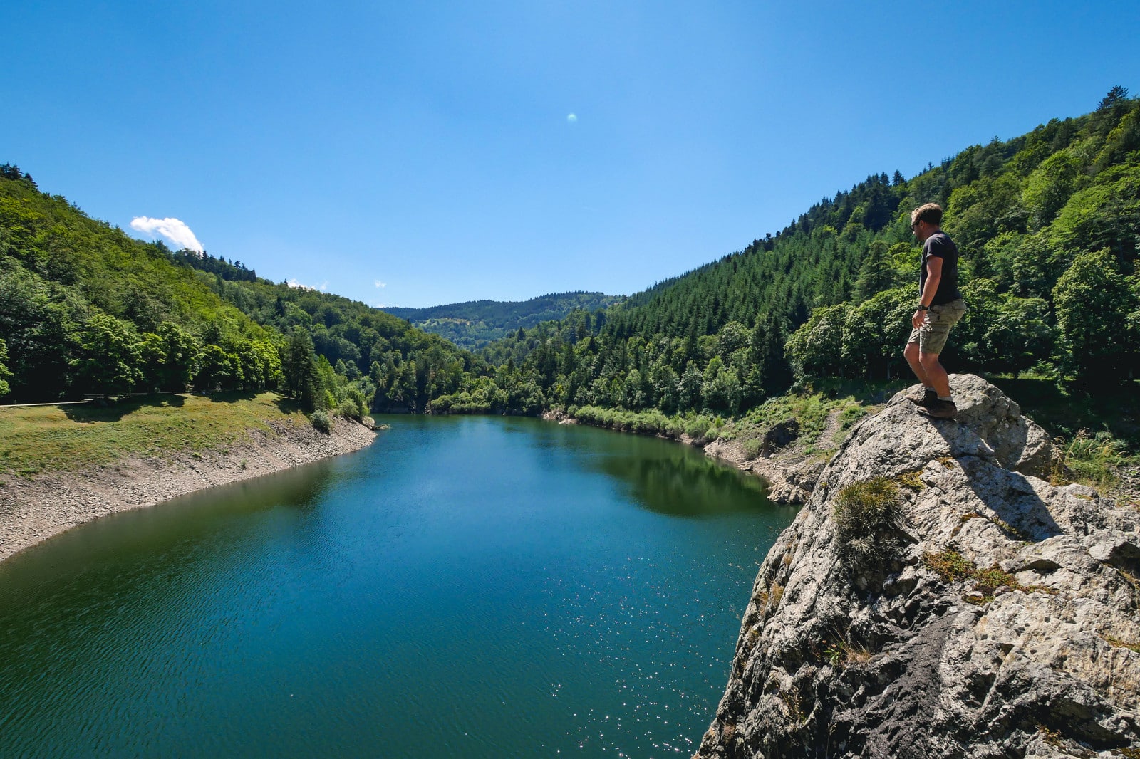 Homme debout sur un rocher surplombant un lac paisible, entouré de collines verdoyantes et de forêts, dans un paysage naturel du Pilat. L'eau calme et le ciel ensoleillé créent une atmosphère sereine et propice à l'évasion.