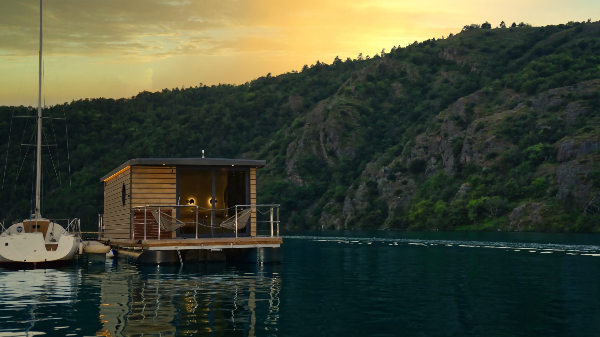 Maison flottante en bois sur une eau calme avec un voilier à côté, entourée de collines verdoyantes et éclairée par un coucher de soleil doré.