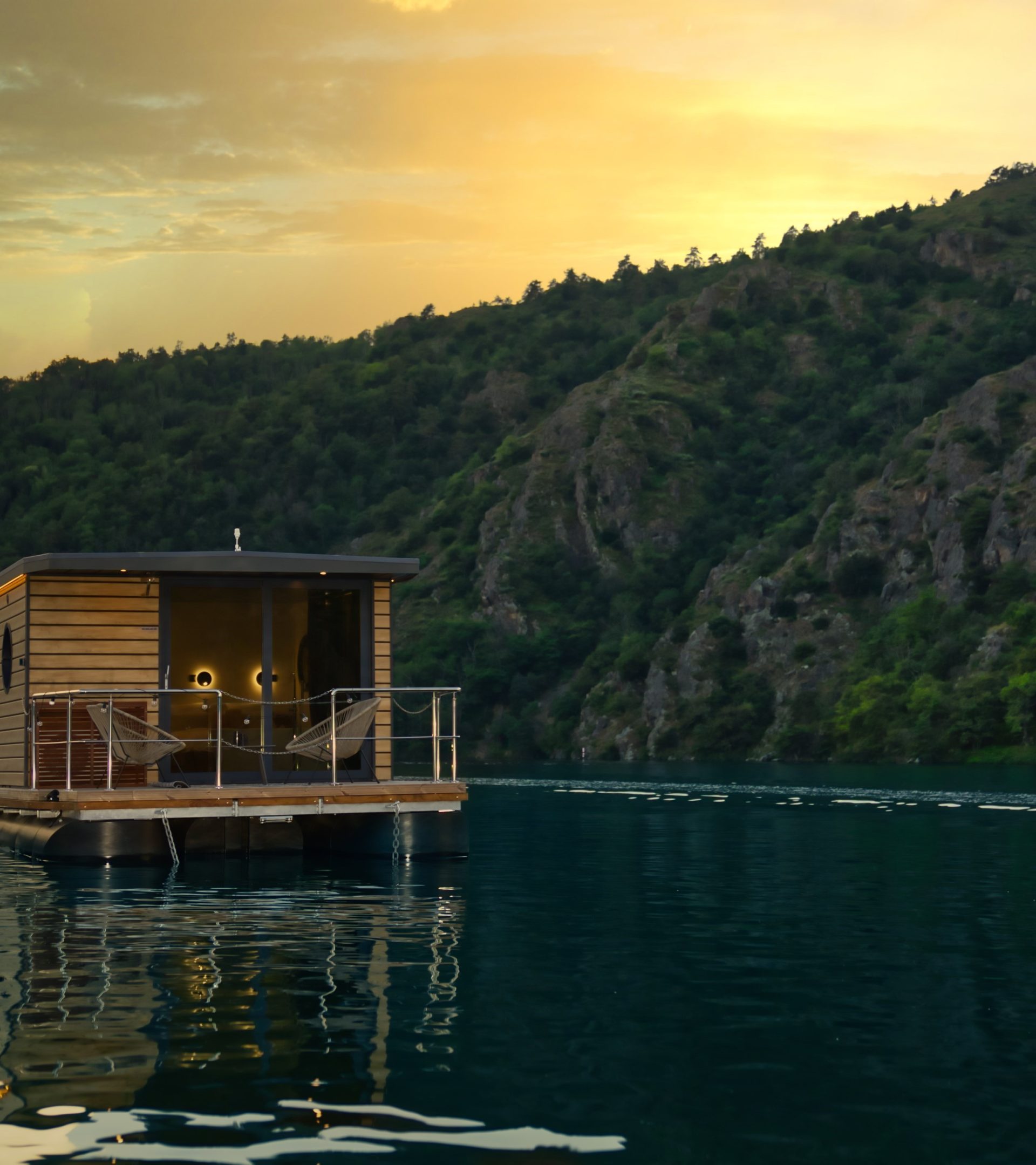 Maison flottante en bois sur une eau calme avec un voilier à côté, entourée de collines verdoyantes et éclairée par un coucher de soleil doré.
