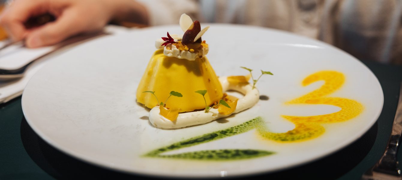 Dessert créatif avec mousse jaune, fruits secs et fleurs comestibles, sur une assiette décorée de sauces colorées.