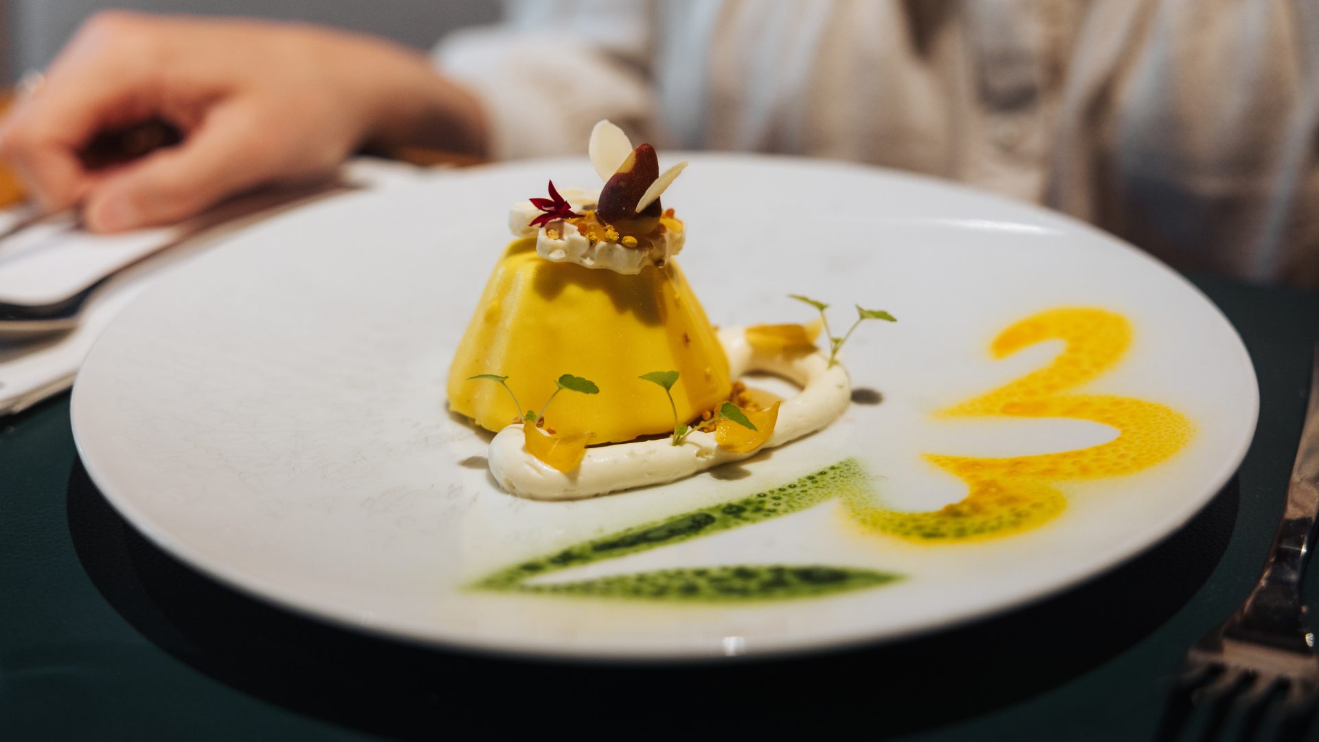 Dessert créatif avec mousse jaune, fruits secs et fleurs comestibles, sur une assiette décorée de sauces colorées.