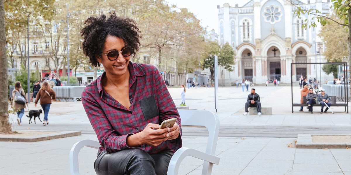 Une personne assise sur une chaise design blanche sur la place Jean-Jaurès à Saint-Étienne, souriante et regardant son téléphone. En arrière-plan, on distingue la façade de l’église Saint-Louis et plusieurs passants profitant de la place animée.