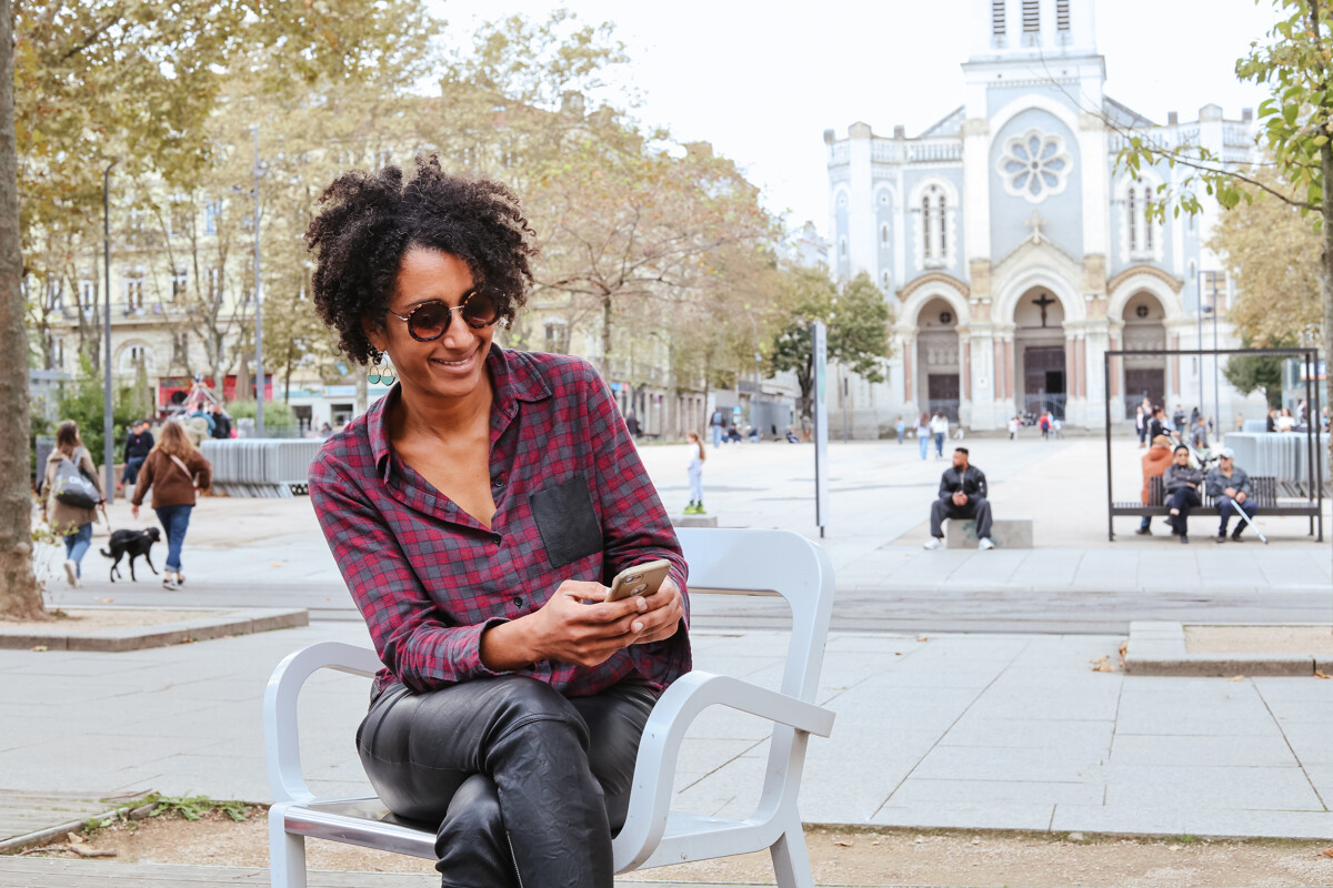 Une personne assise sur une chaise design blanche sur la place Jean-Jaurès à Saint-Étienne, souriante et regardant son téléphone. En arrière-plan, on distingue la façade de l’église Saint-Louis et plusieurs passants profitant de la place animée.