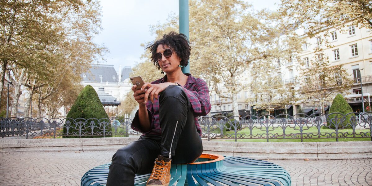 Une personne assise sur un banc design circulaire bleu-vert de la place Jean-Jaurès à Saint-Étienne, utilisant son téléphone portable pour scroller sur les réseaux sociaux. Elle porte une chemise à carreaux, un pantalon noir et des chaussures montantes marron. En arrière-plan, on distingue les arbres d’automne, un kiosque et les façades claires des bâtiments de la place.