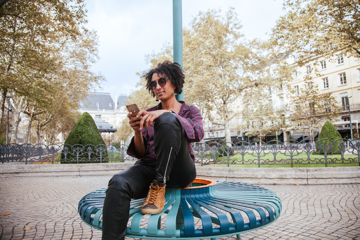 Une personne assise sur un banc design circulaire bleu-vert de la place Jean-Jaurès à Saint-Étienne, utilisant son téléphone portable pour scroller sur les réseaux sociaux. Elle porte une chemise à carreaux, un pantalon noir et des chaussures montantes marron. En arrière-plan, on distingue les arbres d’automne, un kiosque et les façades claires des bâtiments de la place.